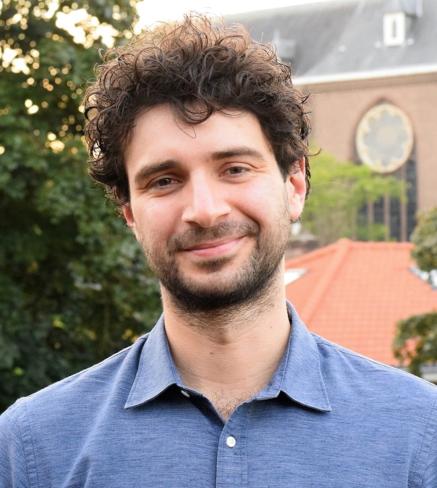 Man van gemiddelde leeftijd met middellang krullend, bruin haar met een blauwe blouse aan. Hij staat buiten in de stad