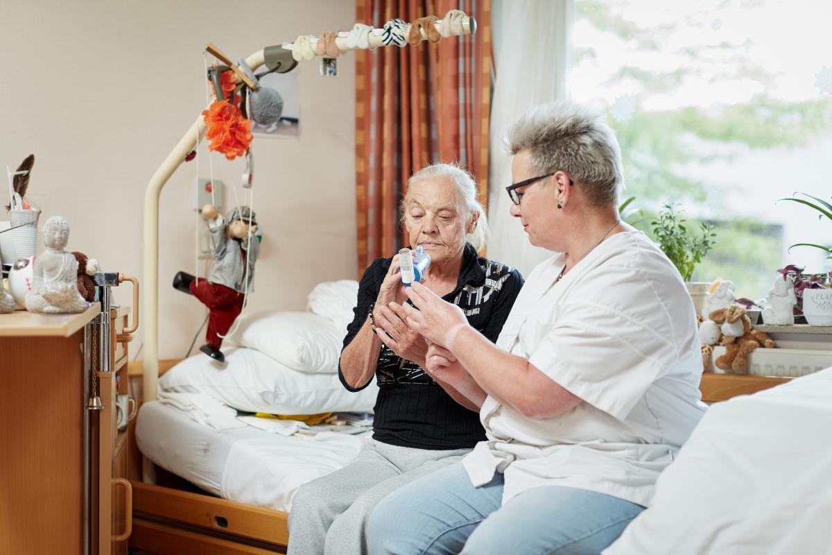 Verpleegkundige zit met cliënt op bed en helpt haar met een inhalator