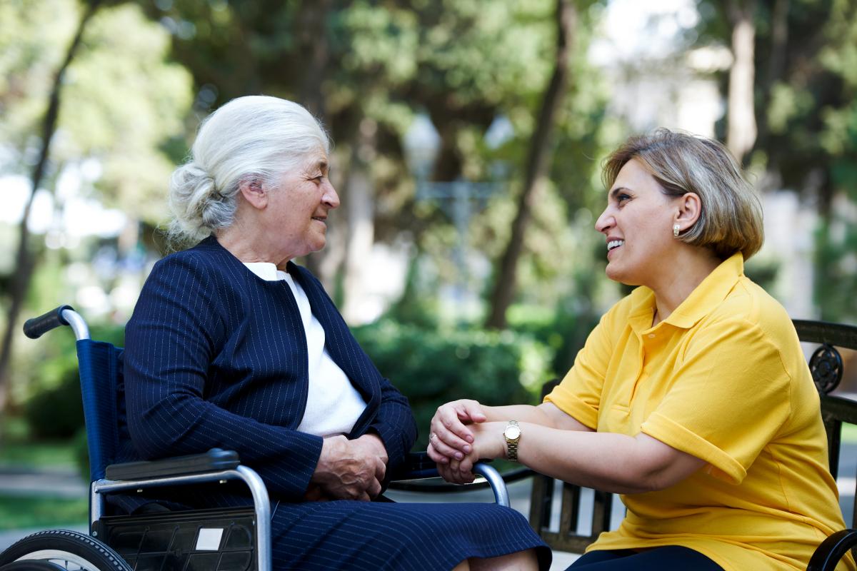 Oudere vrouw in rolstoel met Parkinson in gesprek met naaste die naast haar op een bankje zit in een park